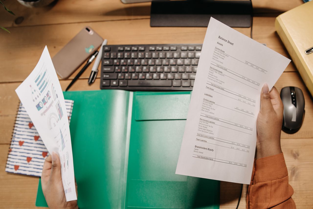 Hands holding and reviewing balance sheets over a desk with a keyboard and stationery.